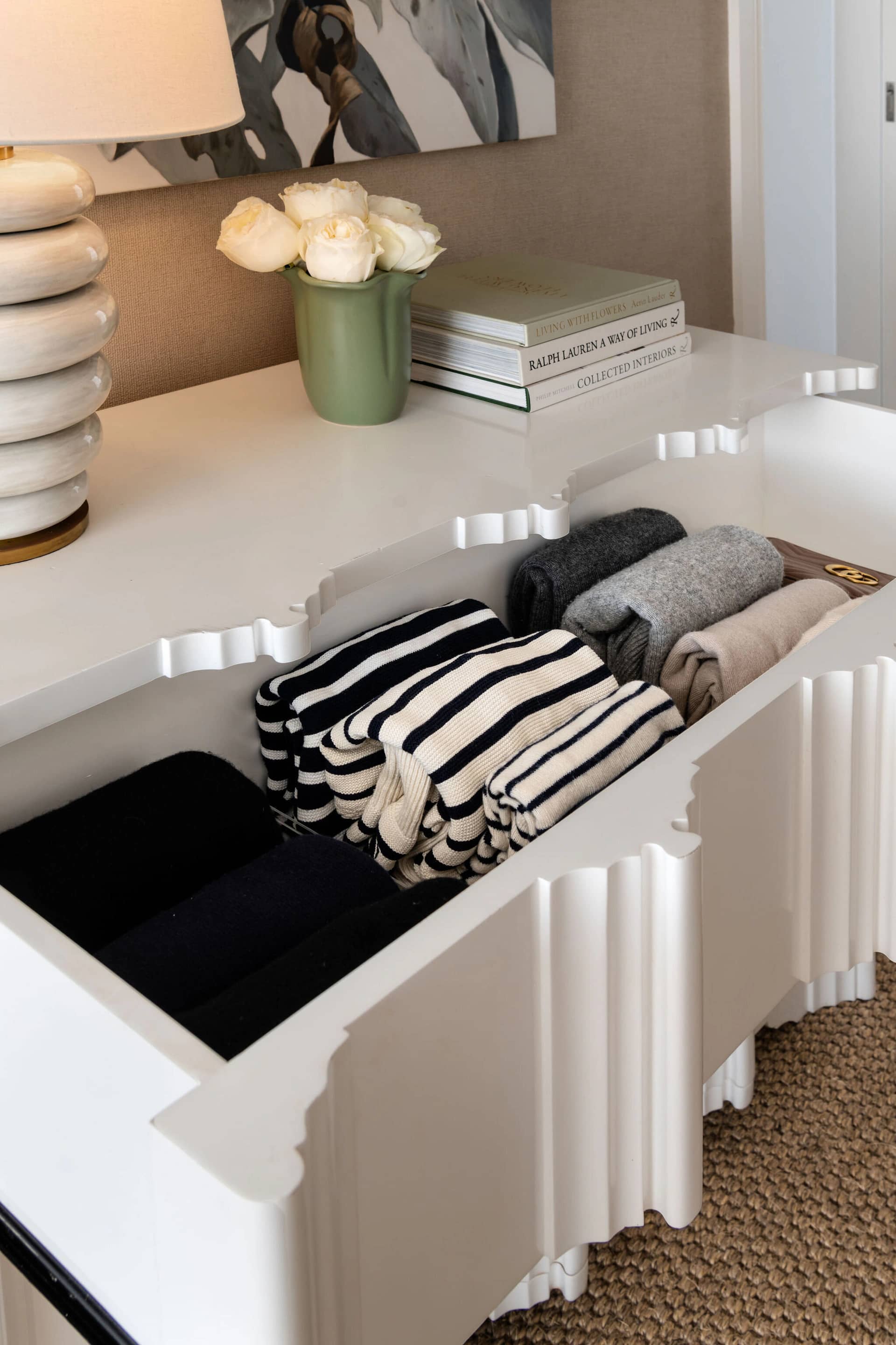 A dresser top with folded knits, books and a vase of roses